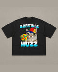 Black heavyweight tee reading GREETINGS HUZZ in blue bubble lettering around a cat wearing a propeller cap and holding a rainbow lollipop