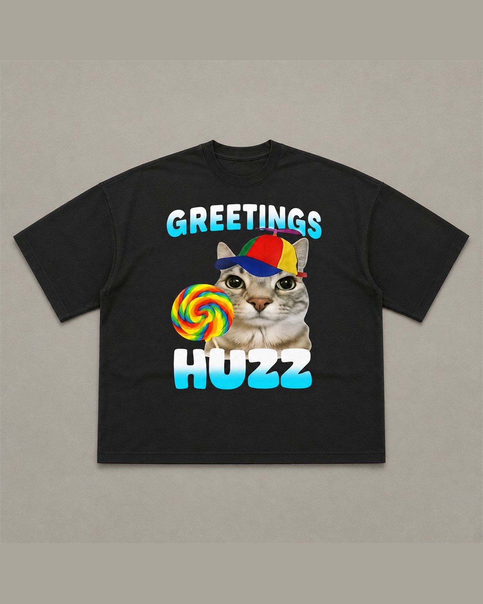 Black heavyweight tee reading GREETINGS HUZZ in blue bubble lettering around a cat wearing a propeller cap and holding a rainbow lollipop